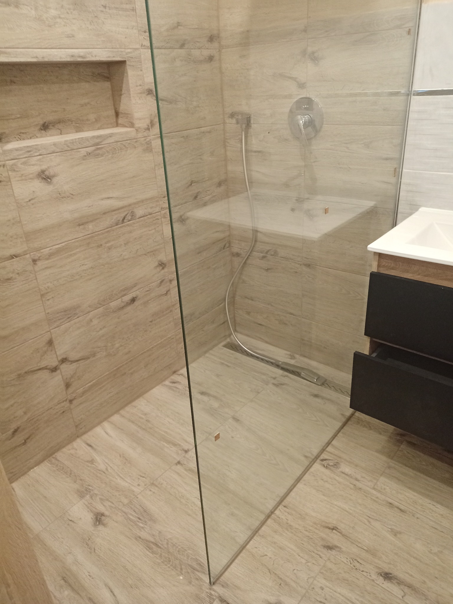 Nowoczesna kabina prysznicowa typu walk-in z odpływem liniowym, wykończona płytkami imitującymi drewno i czarną szafką pod umywalką, z wnęką w ścianie.