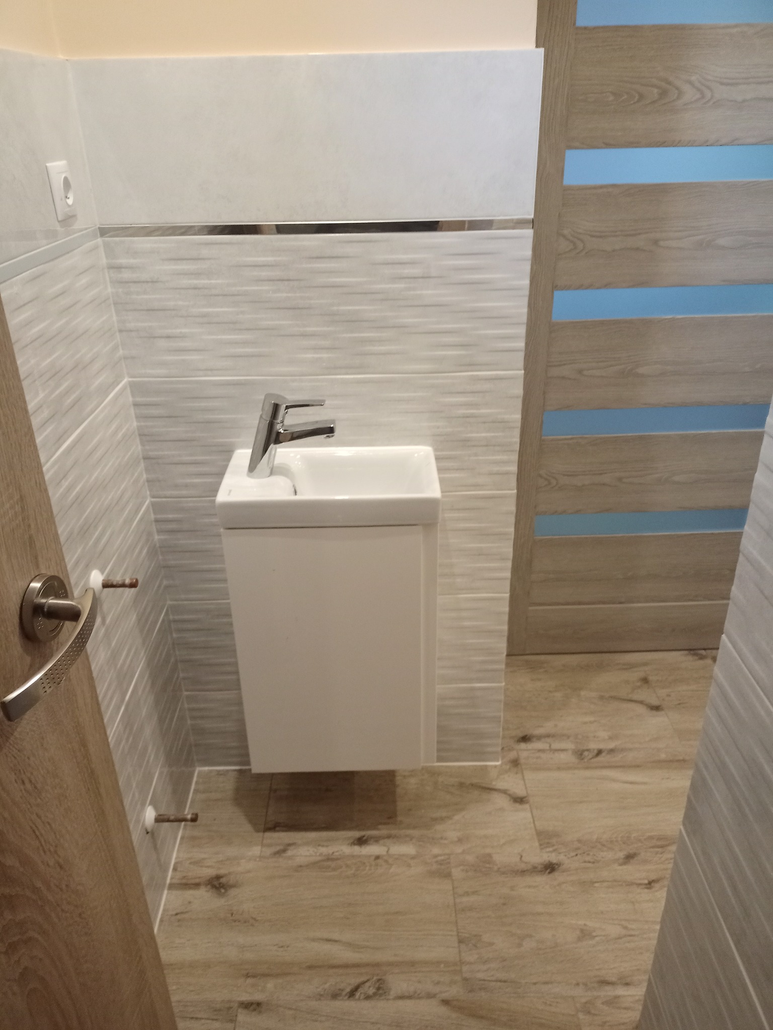 Nowoczesna, mała łazienka z wiszącą umywalką i baterią, wykończona płytkami imitującymi drewno i drzwiami z niebieskimi wstawkami.