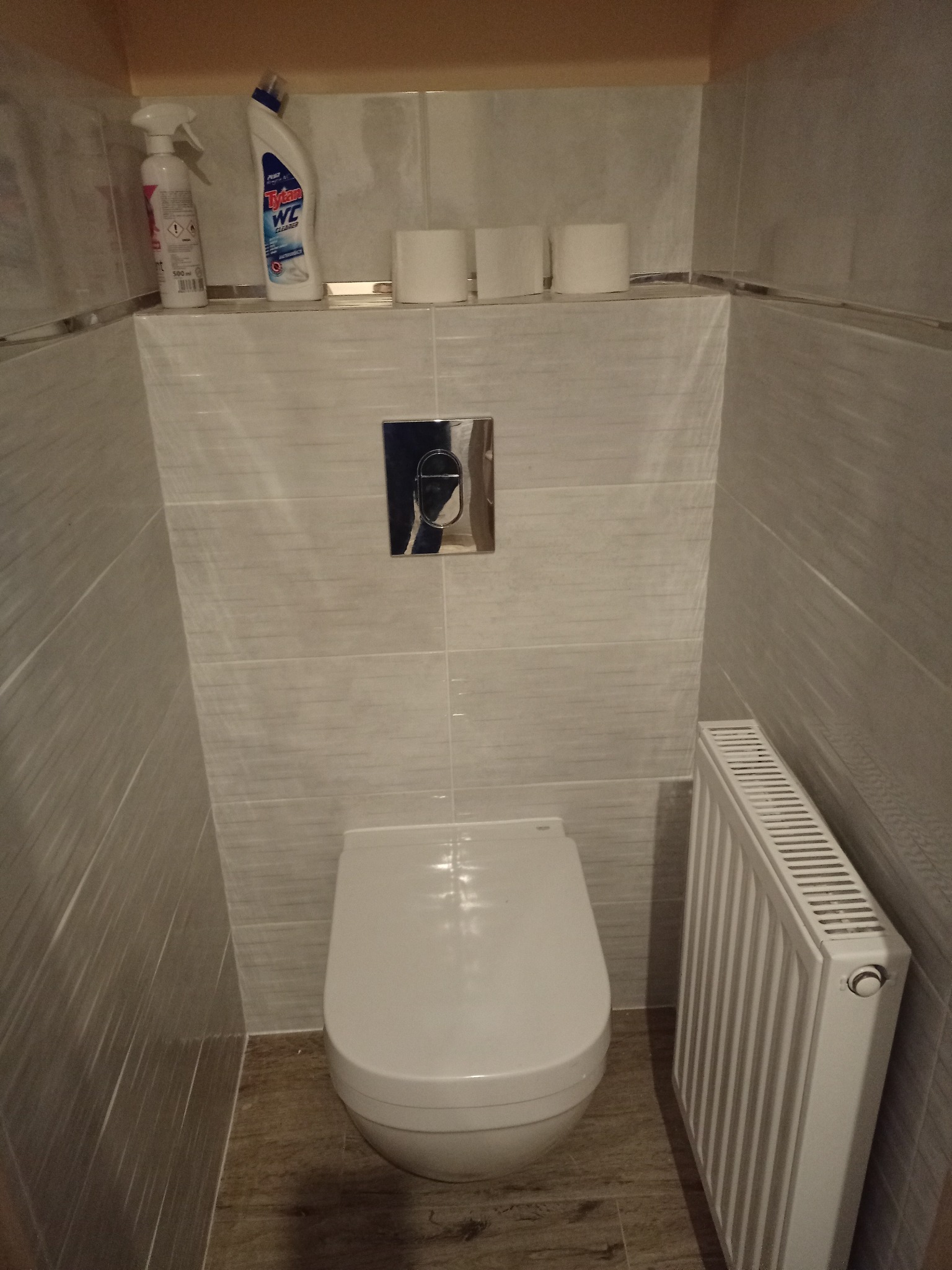 Wykończona toaleta z podwieszaną muszlą, grzejnikiem panelowym i półką nad stelażem, na której stoją środki czystości i zapas papieru toaletowego.