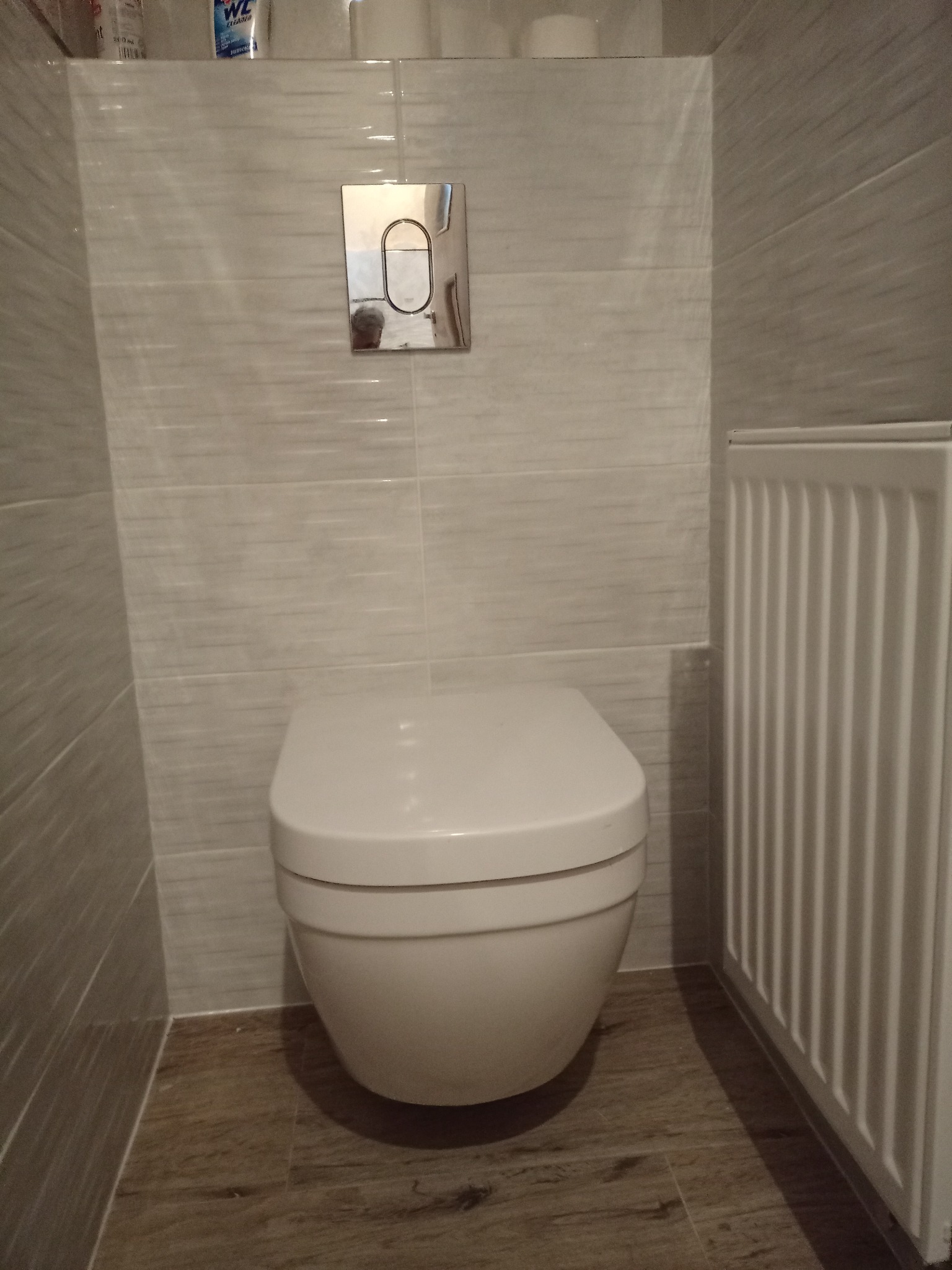 Wisząca toaleta z chromowanym przyciskiem spłukującym na tle płytek imitujących drewno i grzejnika żeberkowego.