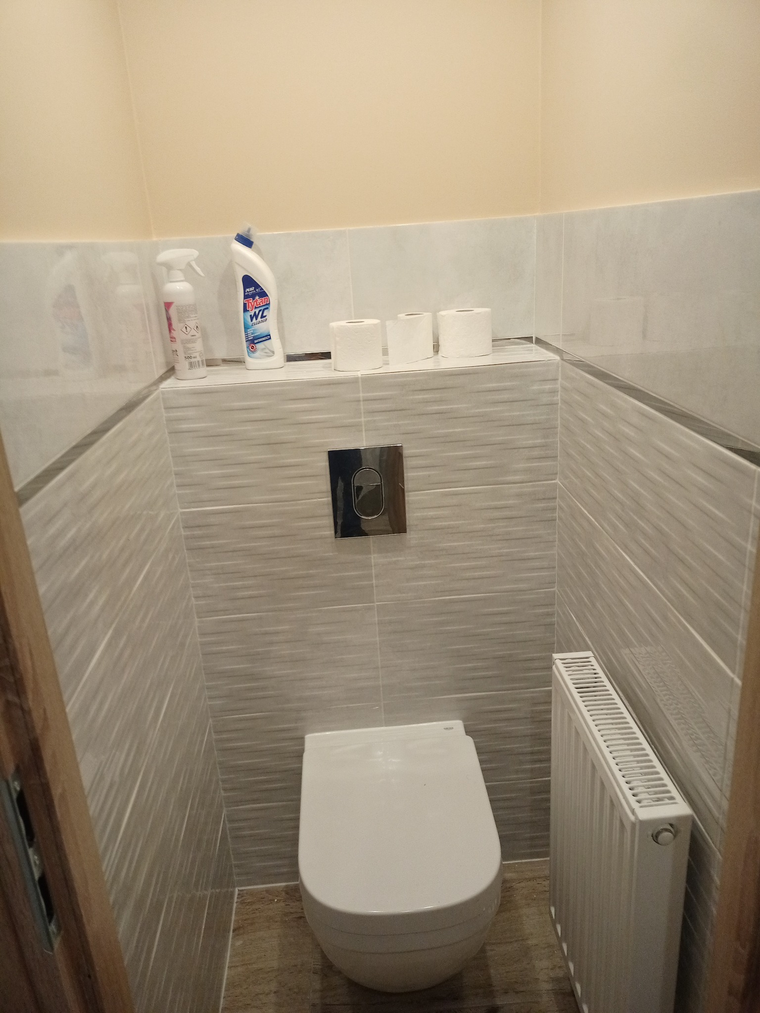 Mała, wykafelkowana toaleta z wiszącą muszlą klozetową, grzejnikiem panelowym i środkami czystości na półce.