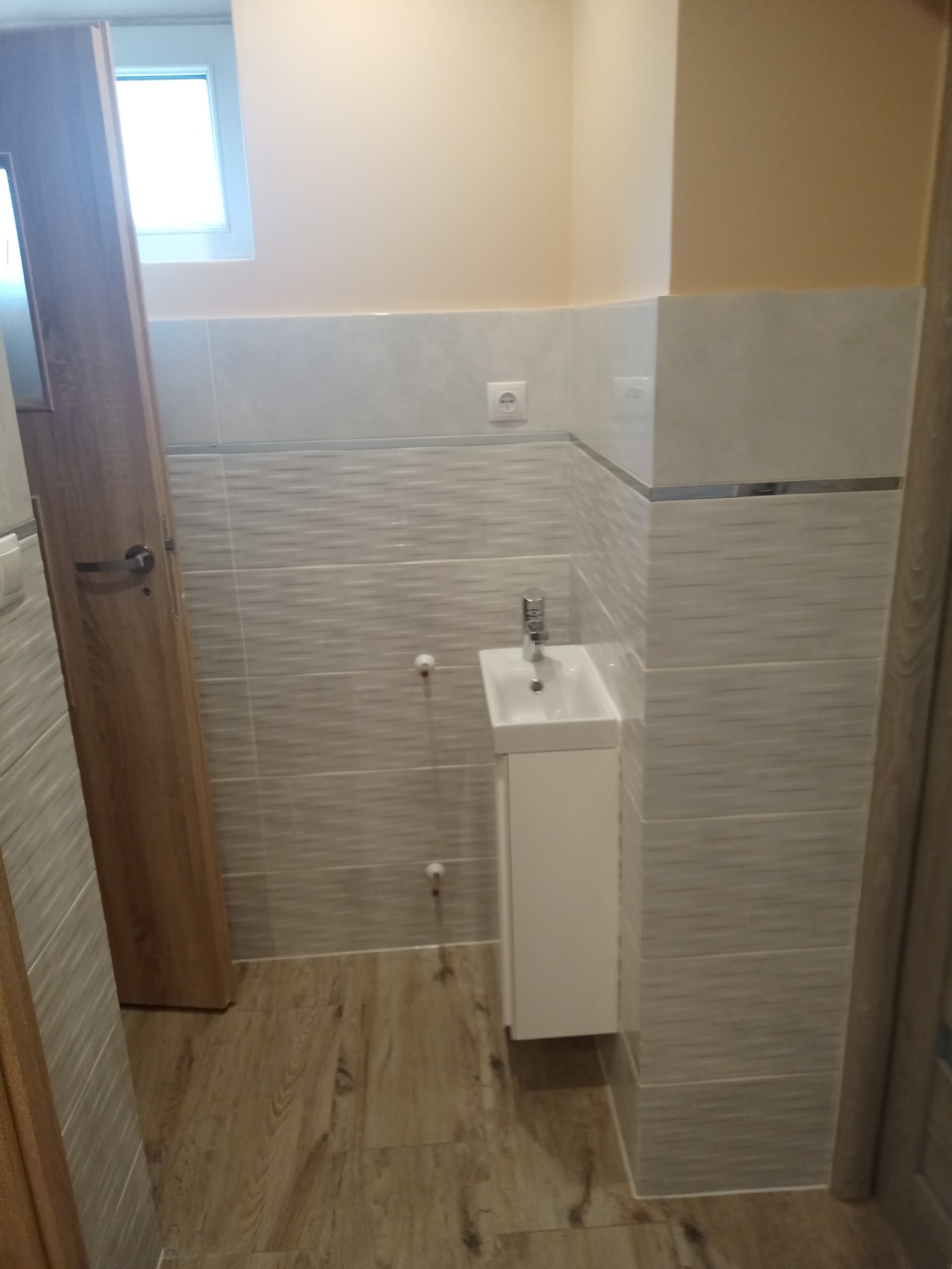 Nowoczesna łazienka z umywalką na szafce, wykończona płytkami imitującymi drewno i jasnymi płytkami na ścianach, z widocznymi elementami instalacji wodnej.