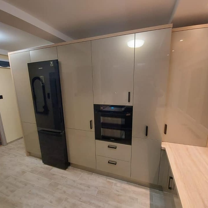 Nowoczesna, beżowa zabudowa kuchenna z czarną lodówką i piekarnikiem, minimalistyczne uchwyty, jasna podłoga, ciepłe oświetlenie.