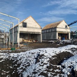 Piotr Mroczkowski Ltd - Dwa budynki w stanie surowym otwartym z konstrukcją dachu, jeden z metalowym szkieletem dobudowy, teren częściowo pokryty śniegiem, w tle las i błękitne niebo.