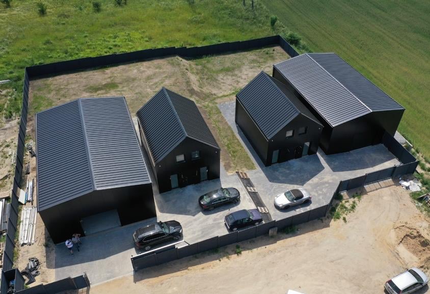 Kompleks nowoczesnych, czarnych budynków z blachy trapezowej, widok z lotu ptaka; zaparkowane samochody osobowe, szara kostka brukowa na podjeździe.