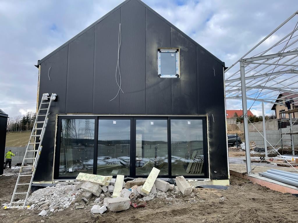 Rozbudowa domu: Czarna elewacja domu z dużymi przesuwnymi oknami, widoczne rusztowanie i gruz budowlany przed budynkiem, drabina oparta o ścianę.