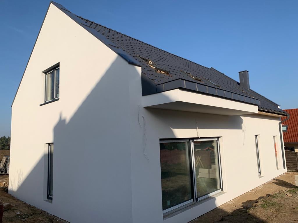 Nowoczesny, biały dom jednorodzinny z płaskim dachem i czarną dachówką, widok z zewnątrz na elewację z dużymi przesuwnymi oknami i balkonem.