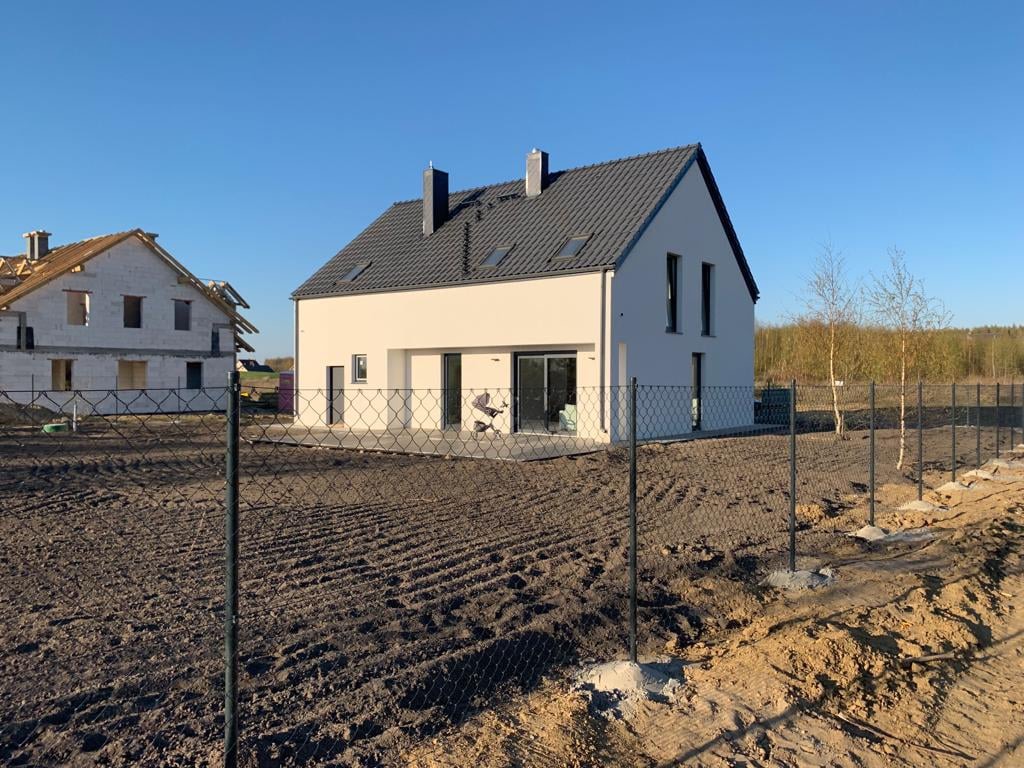 Nowoczesny, biały dom jednorodzinny z ciemną dachówką i dużymi przeszkleniami, widoczny zza ogrodzenia z siatki, obok dom w trakcie budowy.