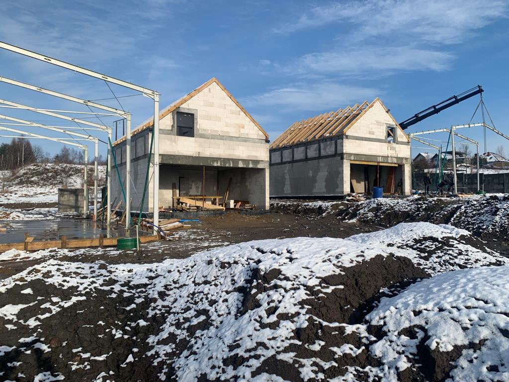 Dwa budynki w stanie surowym otwartym z konstrukcją dachu, jeden z metalowym szkieletem dobudowy, teren częściowo pokryty śniegiem, w tle las i błękitne niebo.