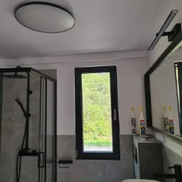 Nowoczesna łazienka z czarnymi akcentami: kabina prysznicowa, okno z widokiem na zieleń, umywalka nablatowa, lampa sufitowa i tuby silikonu na szafce.