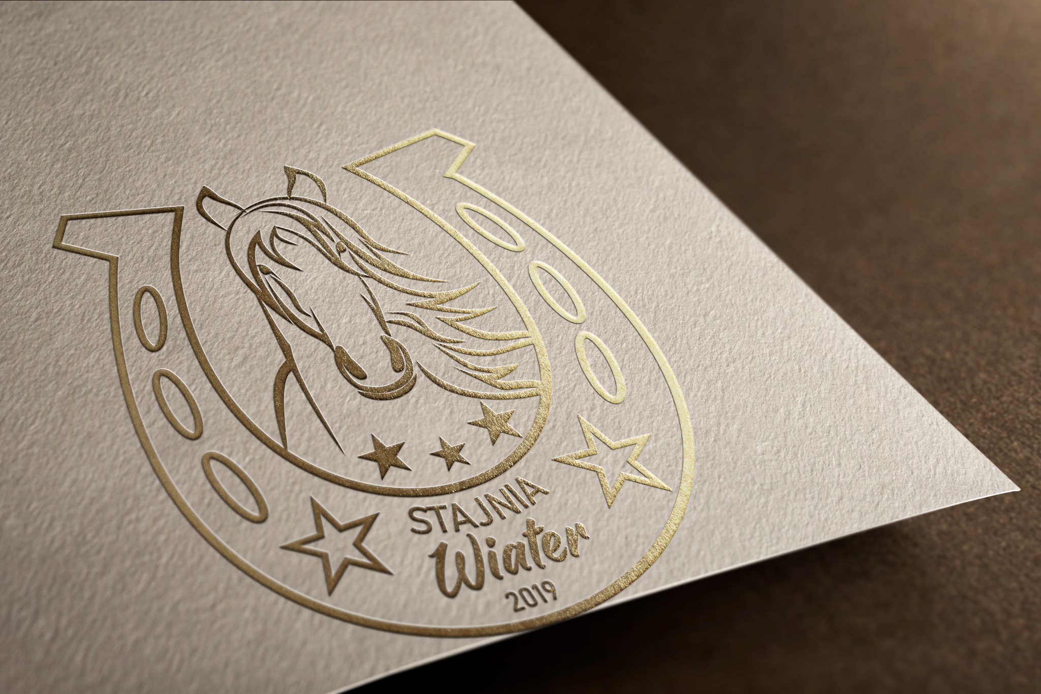 Złote tłoczenie logo 'Stajnia Wiater 2019' z motywem konia i podkowy na kremowym papierze o wyraźnej fakturze, ujęcie pod kątem.