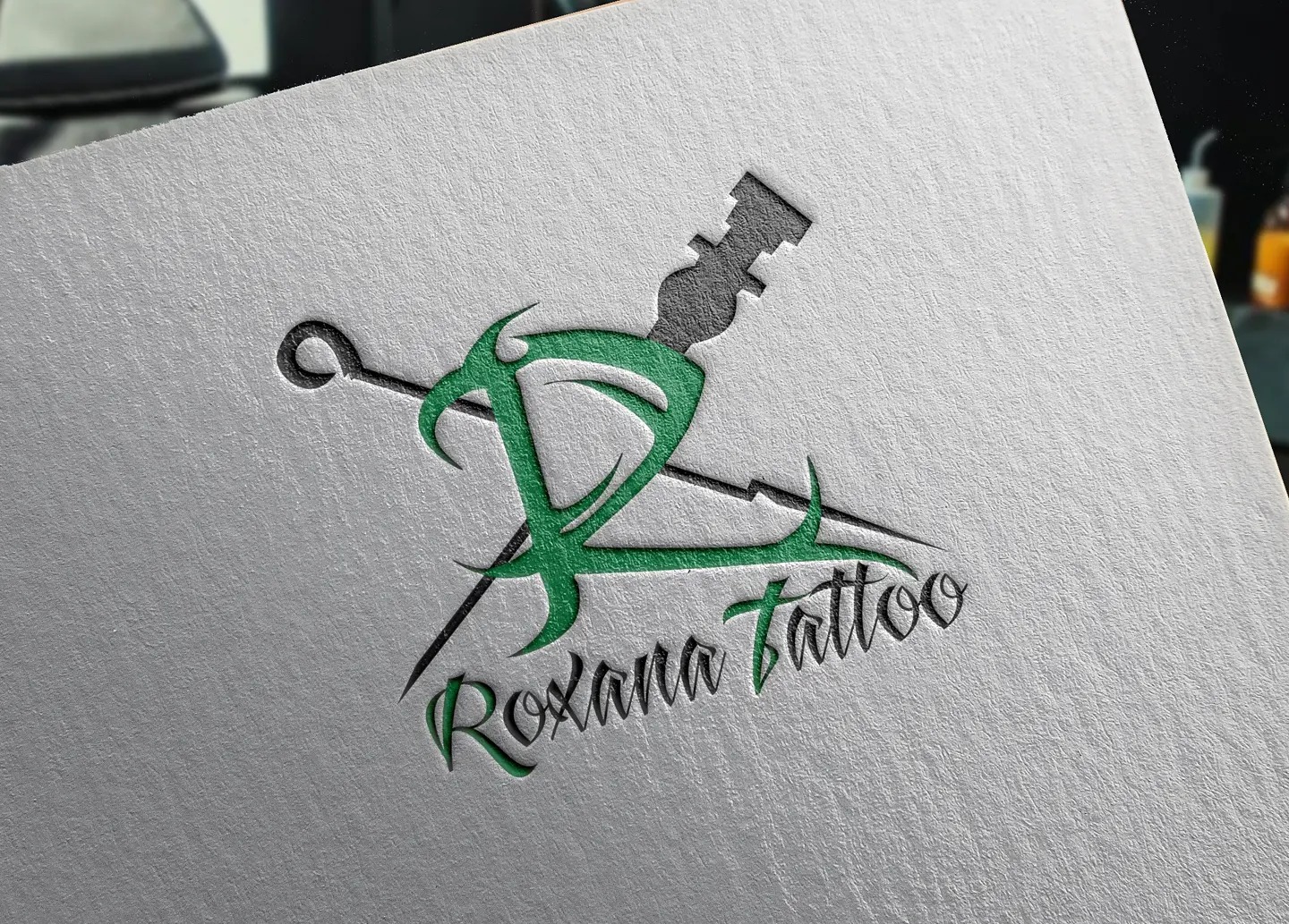 Zielono-czarne logo studia tatuażu 'Roxana Tattoo' z motywem igły do tatuażu na teksturowanym, białym papierze.