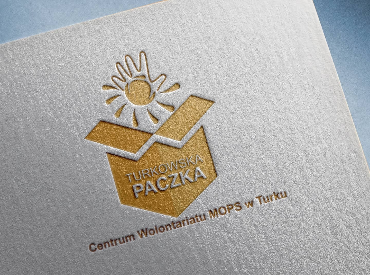Złote logo „Turkowska Paczka Centrum Wolontariatu MOPS w Turku” wytłoczone na białym, teksturowanym papierze.