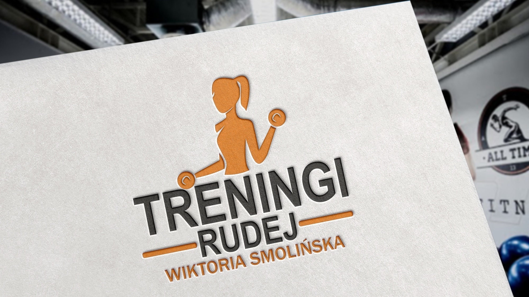 Mockup logo 'Treningi Rudej' Wiktoria Smolińska, z sylwetką kobiety ćwiczącej z hantlami, umieszczony na białym papierze z fakturą, w tle widoczne elementy siłowni.