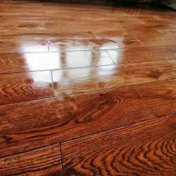 Błyszcząca, świeżo położona podłoga z paneli imitujących drewno, widoczne odbicie światła od okna, ciepła kolorystyka brązu i beżu.