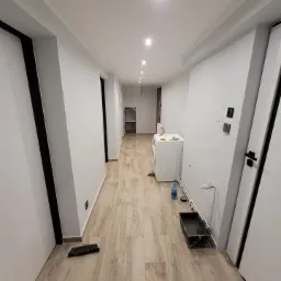 Długi korytarz po remoncie, z nowymi panelami podłogowymi imitującymi drewno, świeżo pomalowanymi ścianami na szaro i białymi drzwiami z czarnymi ościeżnicami; widoczna pralka i akcesoria malarskie.