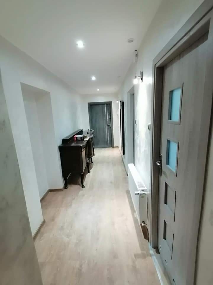 Wykończony korytarz z panelami podłogowymi imitującymi drewno, białe ściany i sufit z oświetleniem punktowym, ciemny kredens przy ścianie i szare drzwi z przeszkleniami.