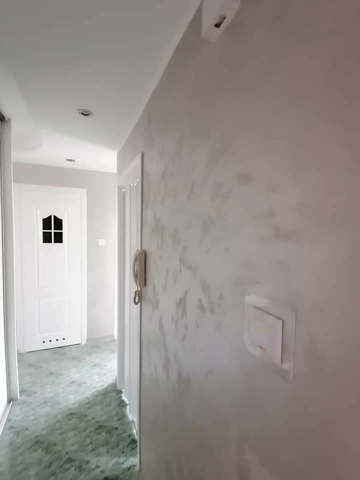 Korytarz po odświeżeniu, ściana z efektem dekoracyjnym w odcieniach szarości, widoczny włącznik światła i domofon, białe drzwi w głębi z czarnymi szybkami, podłoga w odcieniach zieleni.