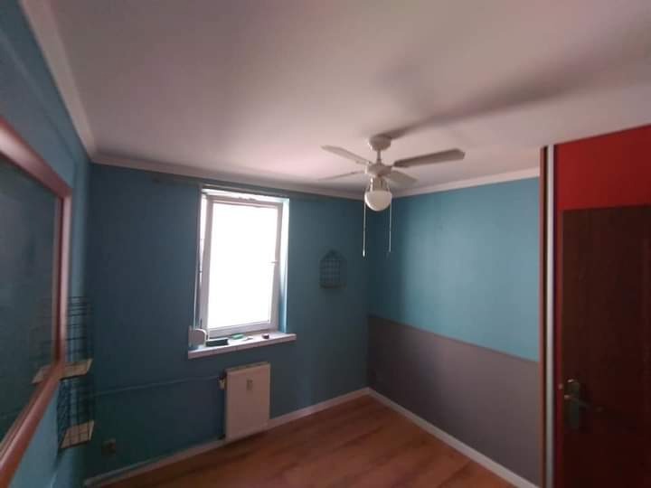 Pomieszczenie po malowaniu ścian na niebiesko z szarym pasem na dole, widoczny wentylator sufitowy i drewniana podłoga.