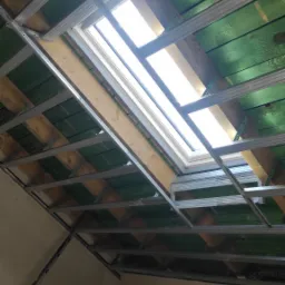Montaż metalowej konstrukcji pod sufitem z oknem dachowym, widoczne zielone deski i narzędzia na podłodze.