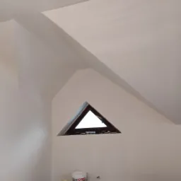 Poddasze po remoncie, białe ściany, trójkątne okno z brązową ramą, widoczny fragment drzwi i pojemnik z masą szpachlową CEKOL.