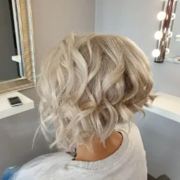 Krótkie, platynowe, kręcone włosy, widok z tyłu, w tle lustro z oświetleniem i fragment blatu.