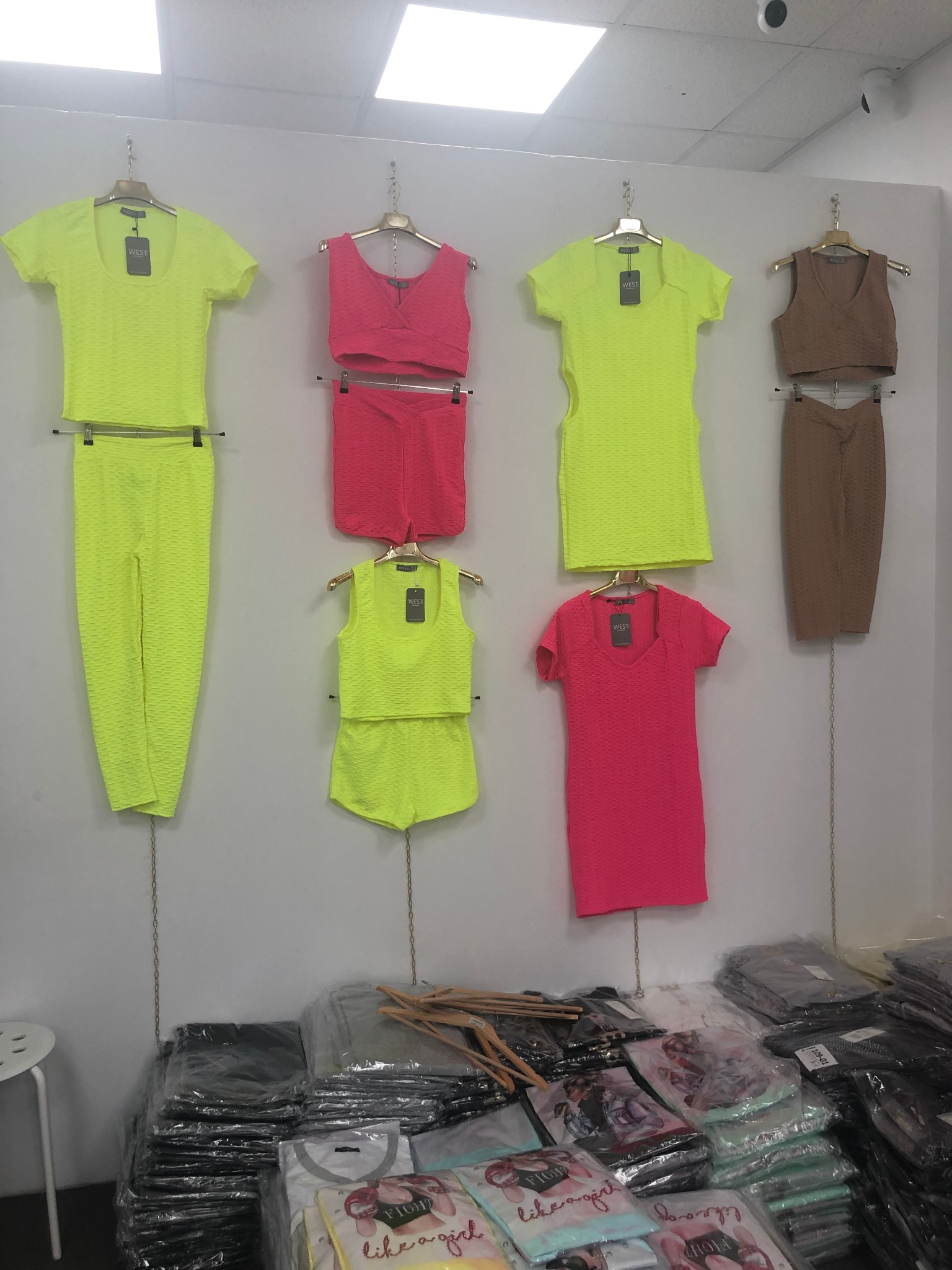 Wystawa ubrań damskich na wieszakach: neonowo-żółty komplet spodni i bluzki, różowy komplet topu i spodenek, neonowo-żółty komplet topu i spodenek, różowa sukienka oraz brązowy komplet topu...