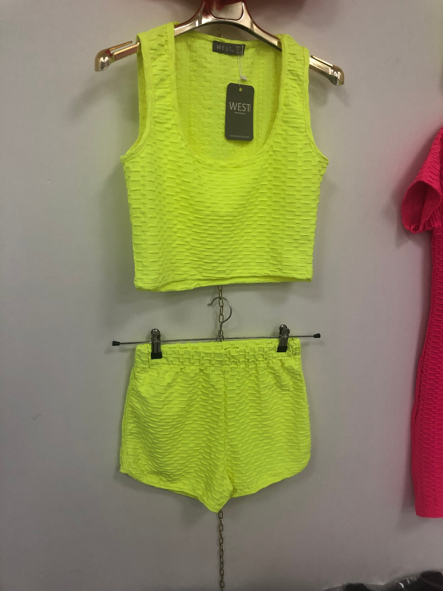 Neonowy, teksturowany komplet sportowy: krótki top i szorty, wiszące na wieszakach na tle szarej ściany. Metka marki WEST widoczna na topie. Po prawej fragment różowej sukienki.