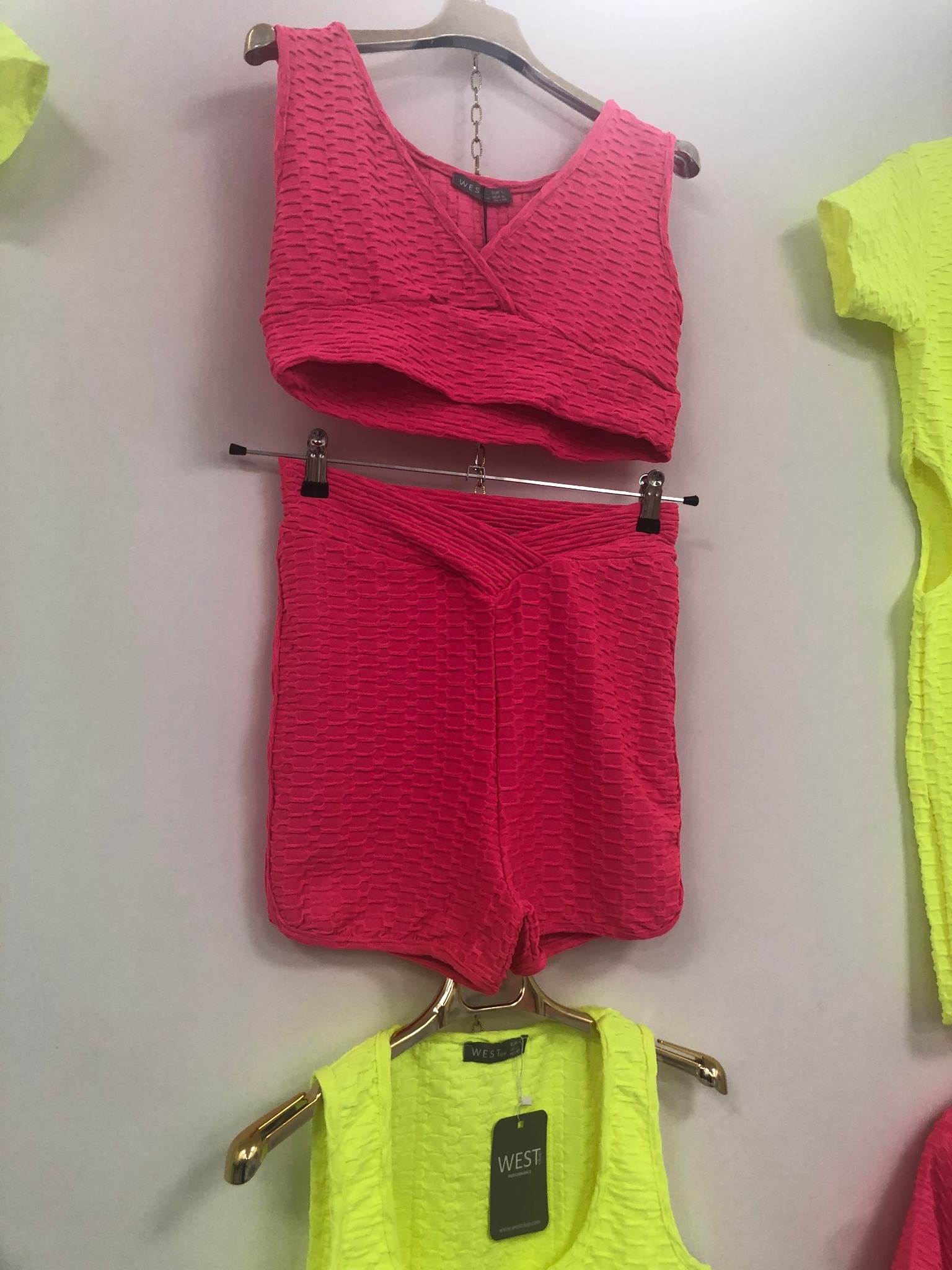 Różowy komplet sportowy (top i szorty) z teksturą wafelkową, wiszący na wieszaku obok neonowej kamizelki z metką marki WEST.