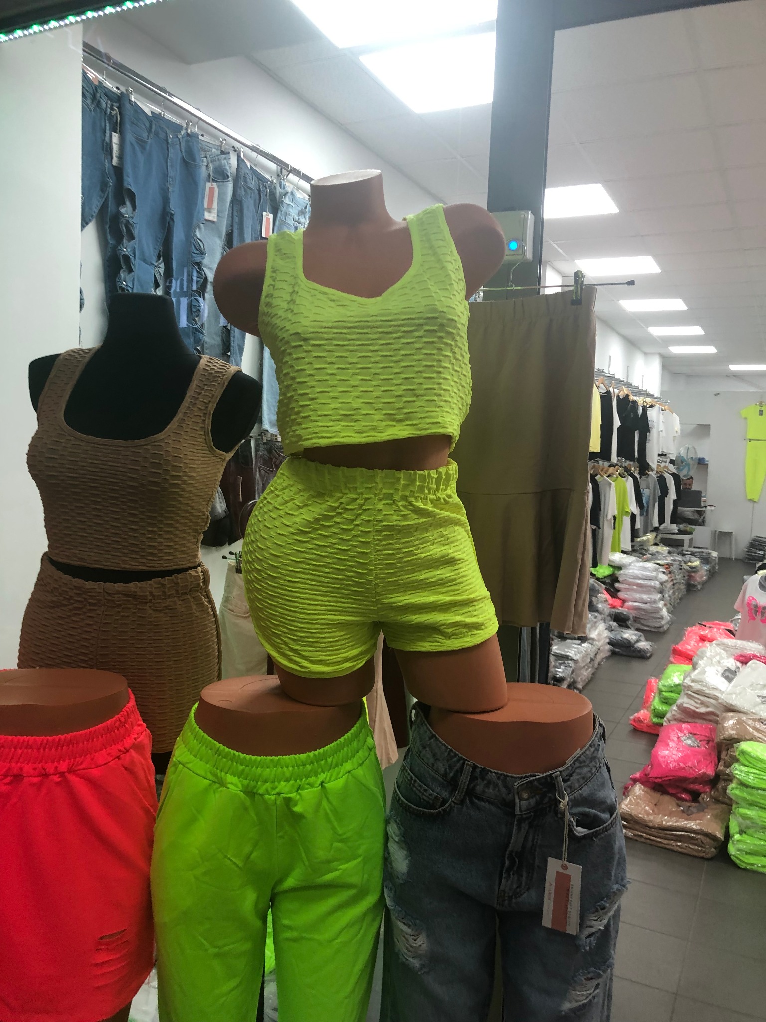 Manekiny prezentują komplety: neonowo-zielony top i szorty z fakturą oraz brązowy komplet. W tle widoczne dżinsy z dziurami i inne ubrania na wieszakach.