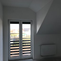 Bartosz Guz - Pokój na poddaszu z oknem balkonowym, grzejnikiem i surowym betonowym podłożem przygotowanym pod wykończenie.