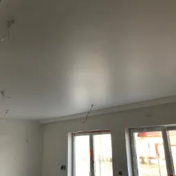 Białe, świeżo wykończone wnętrze z widocznymi przewodami elektrycznymi wystającymi z sufitu, duże okna z widokiem na dom z czerwonym dachem.