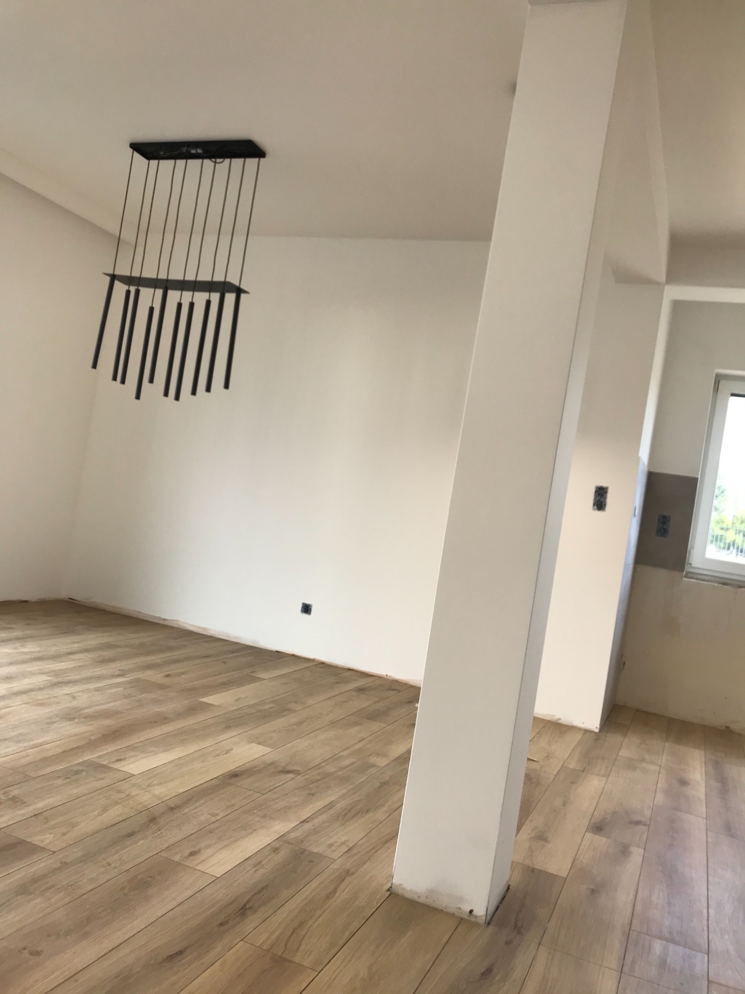 Wnętrze po malowaniu ścian na biało, widoczna drewniana podłoga i nowoczesna lampa wisząca z czarnymi elementami, biały filar w przejściu do innego pomieszczenia.