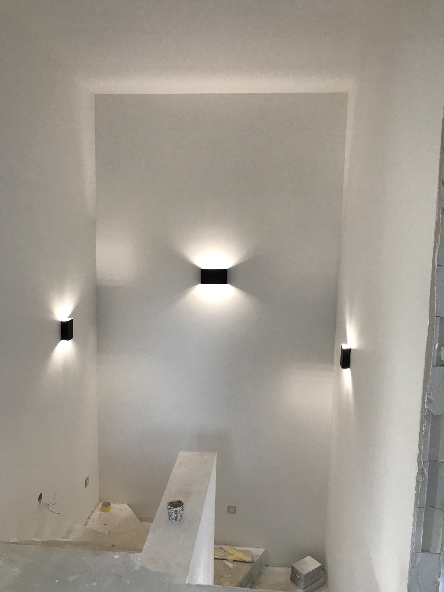 Klatka schodowa z gładzią, oświetlona trzema czarnymi kinkietami, z widocznymi śladami prac wykończeniowych i puszkami po farbie na schodach.