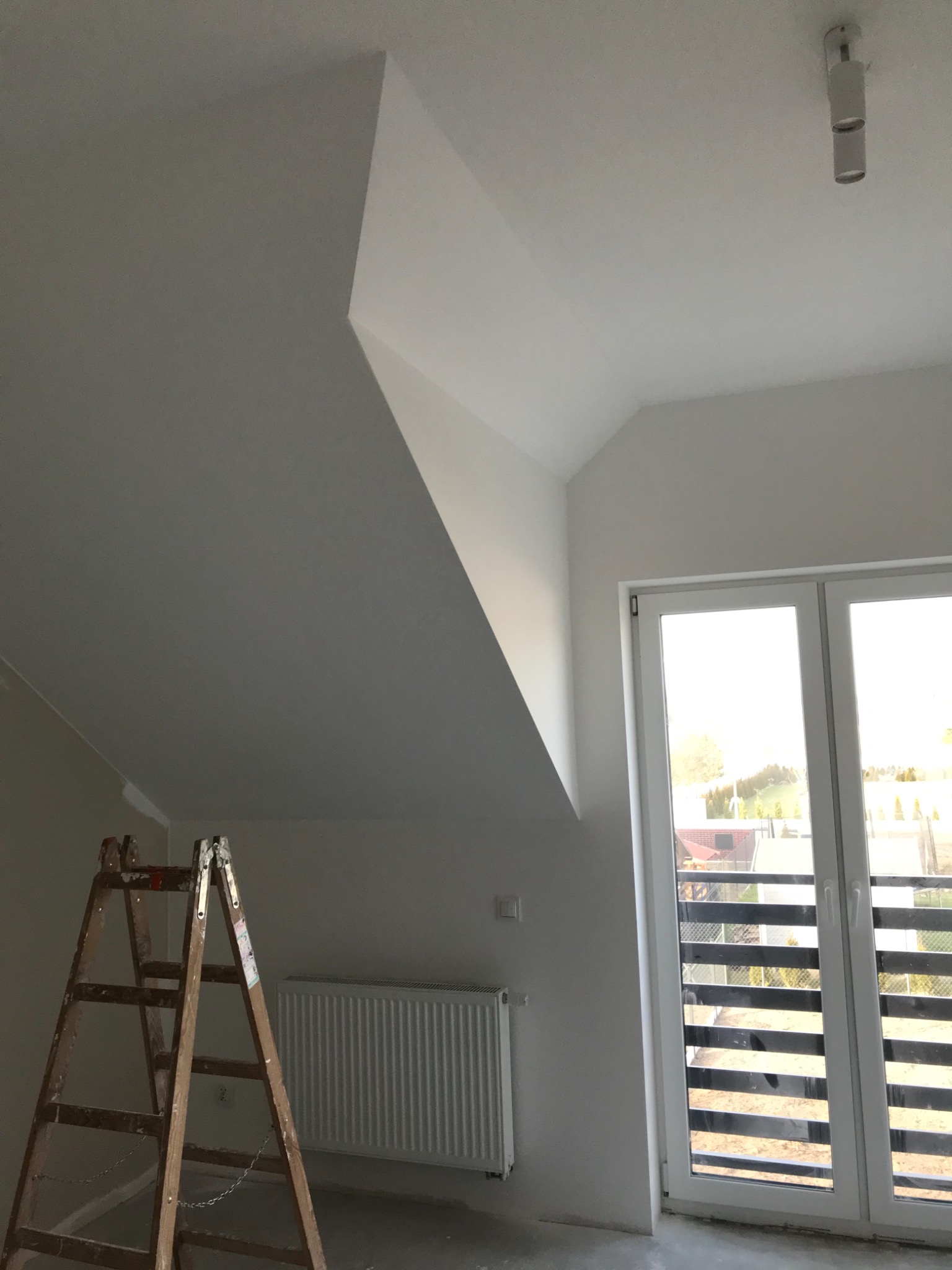 Wnętrze poddasza po świeżym pomalowaniu na biało, widoczna drewniana drabina, grzejnik i okno balkonowe z widokiem na zewnątrz.