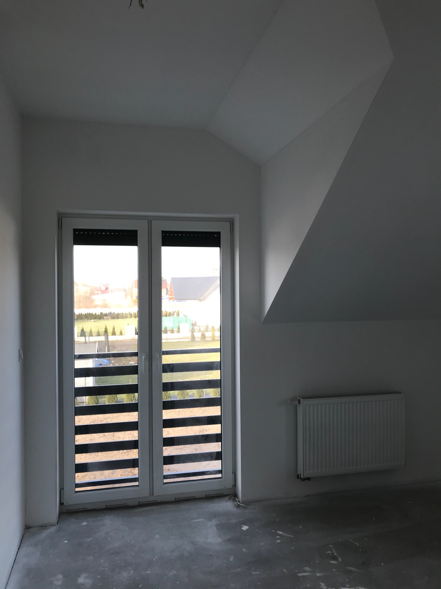 Pokój na poddaszu z oknem balkonowym, grzejnikiem i surowym betonowym podłożem przygotowanym pod wykończenie.