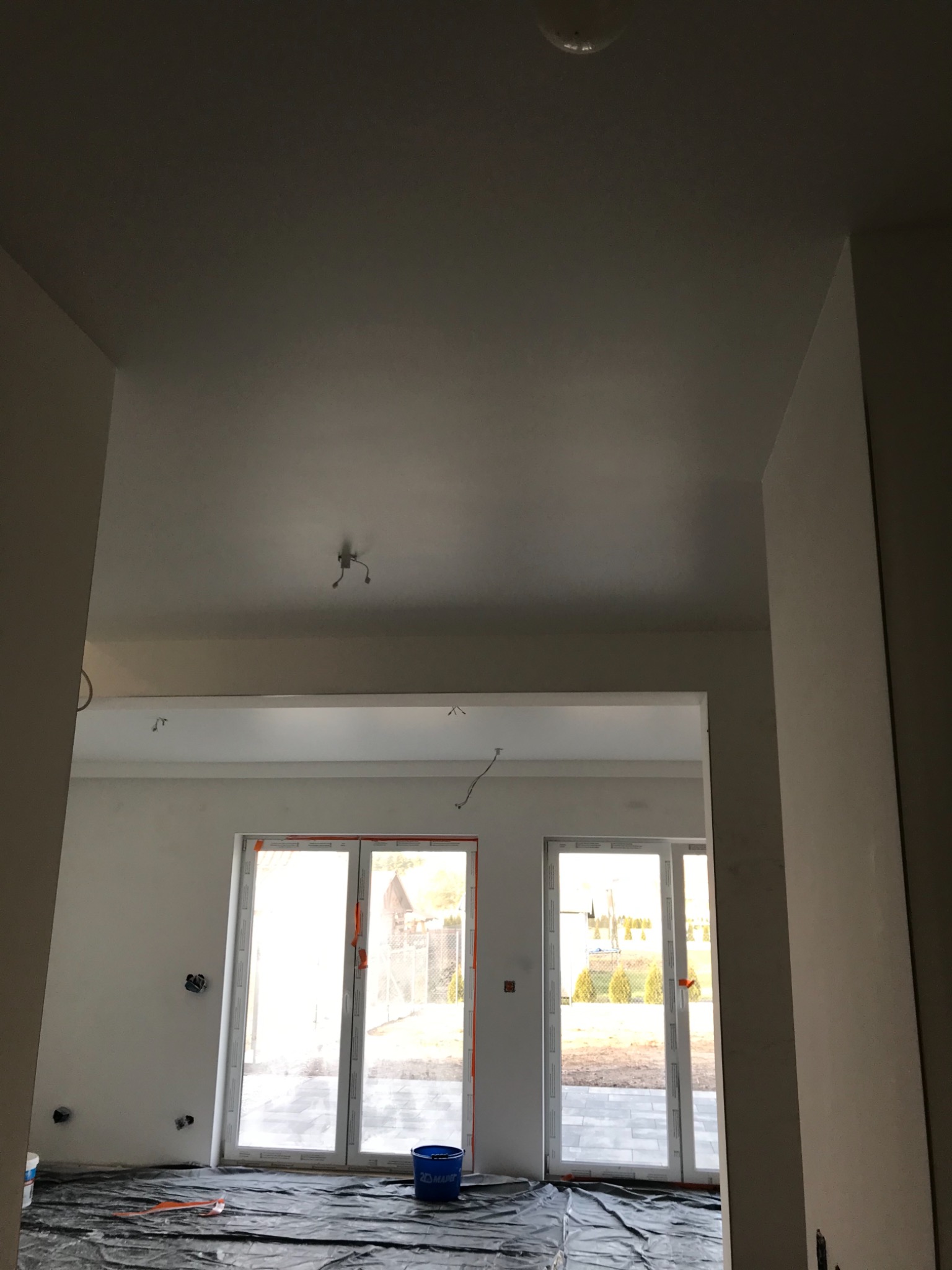 Wnętrze w trakcie remontu: świeżo pomalowane ściany i sufit na biało, zabezpieczona folią podłoga, widoczne puszki elektryczne i niepodłączone lampy sufitowe, błękitne wiadro na podłodze.