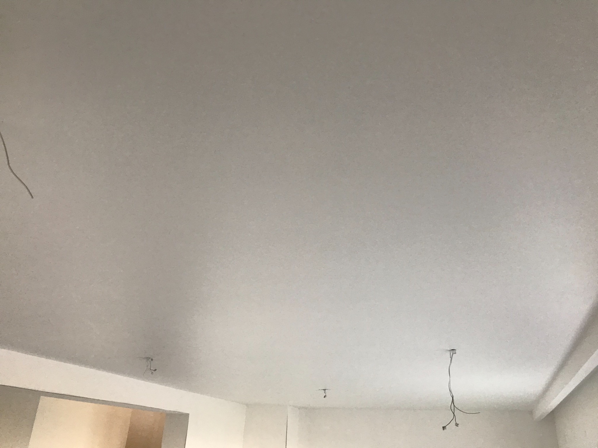 Gładka, biała powierzchnia sufitu po szpachlowaniu i malowaniu, widoczne przewody oświetleniowe wystające z otworów, fragment ściany w tle.