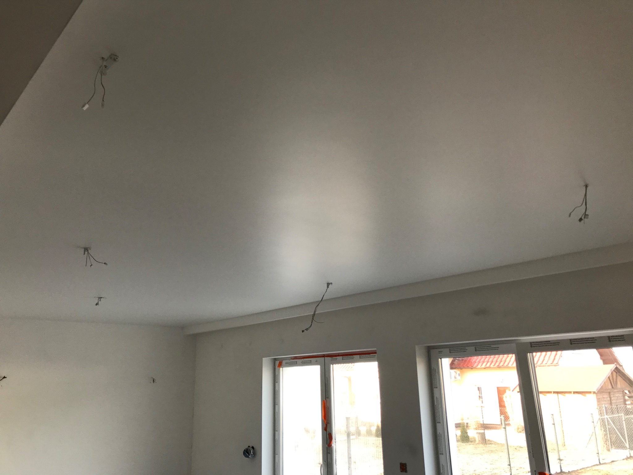 Białe, świeżo wykończone wnętrze z widocznymi przewodami elektrycznymi wystającymi z sufitu, duże okna z widokiem na dom z czerwonym dachem.