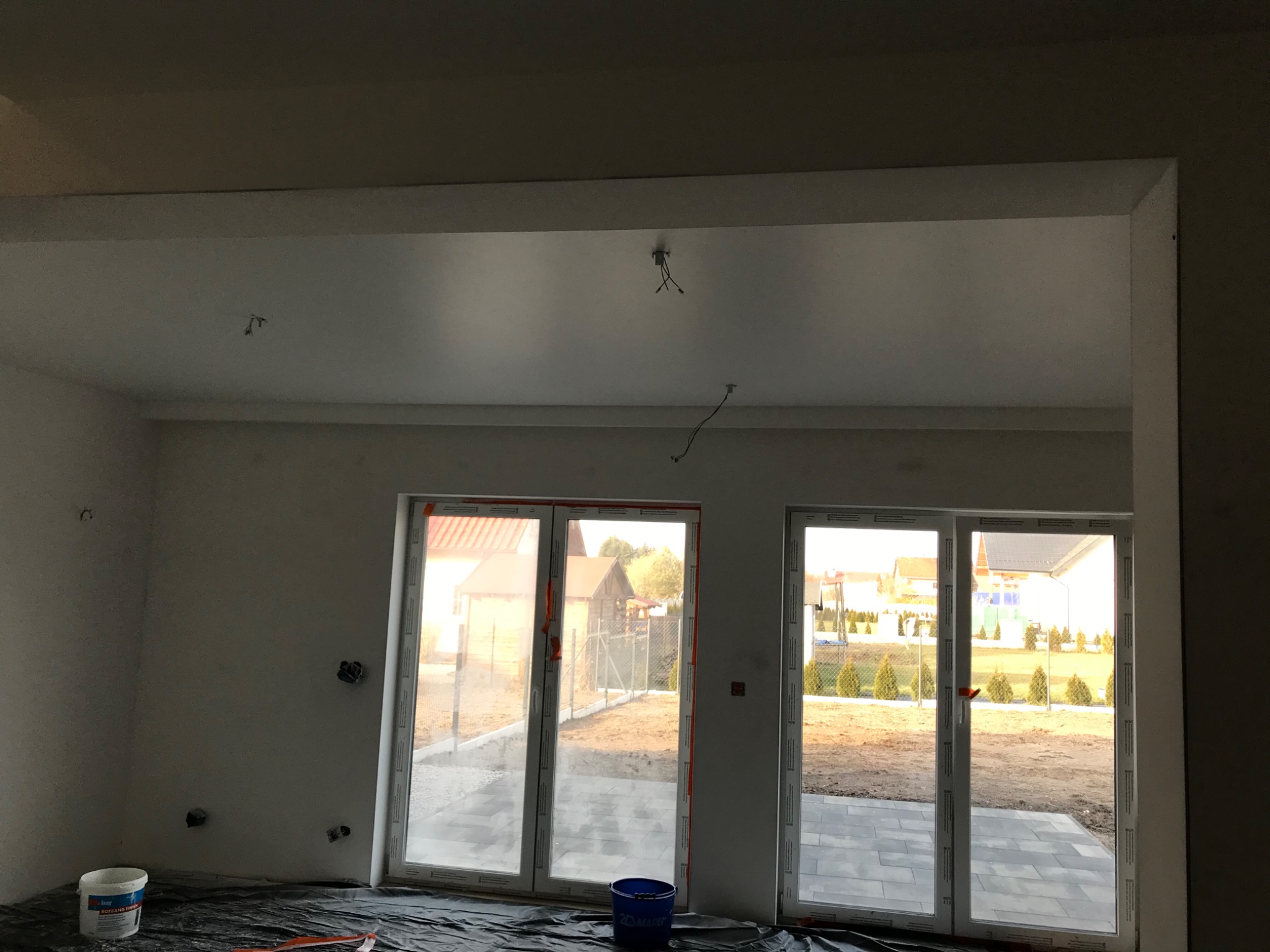 Wnętrze w stanie deweloperskim z przygotowanymi ścianami pod malowanie, zabezpieczona podłoga folią, widoczne puszki elektryczne bez osprzętu, dwa okna balkonowe z widokiem na ogród.