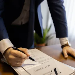 Osoba w garniturze podpisuje dokument 'Akta Sprawy' na drewnianym biurku, widoczny zegarek na nadgarstku i fragment zielonej rośliny w tle.