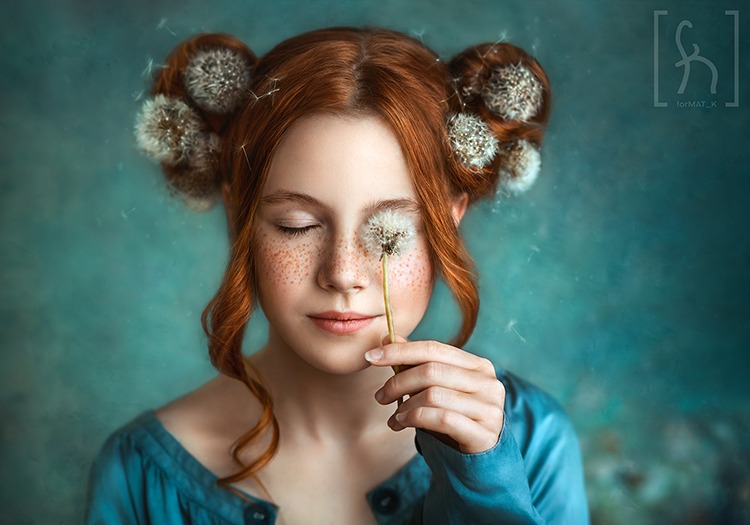 Portret rudej dziewczyny z zamkniętymi oczami, trzymającej dmuchawca przed twarzą, z dmuchawcami we włosach, na rozmytym turkusowym tle.