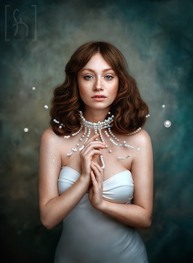 Portret młodej kobiety z rudymi włosami i piegami, ubrana w białą sukienkę bez ramiączek i oryginalny naszyjnik z pereł, na rozmytym, ciemnym tle.