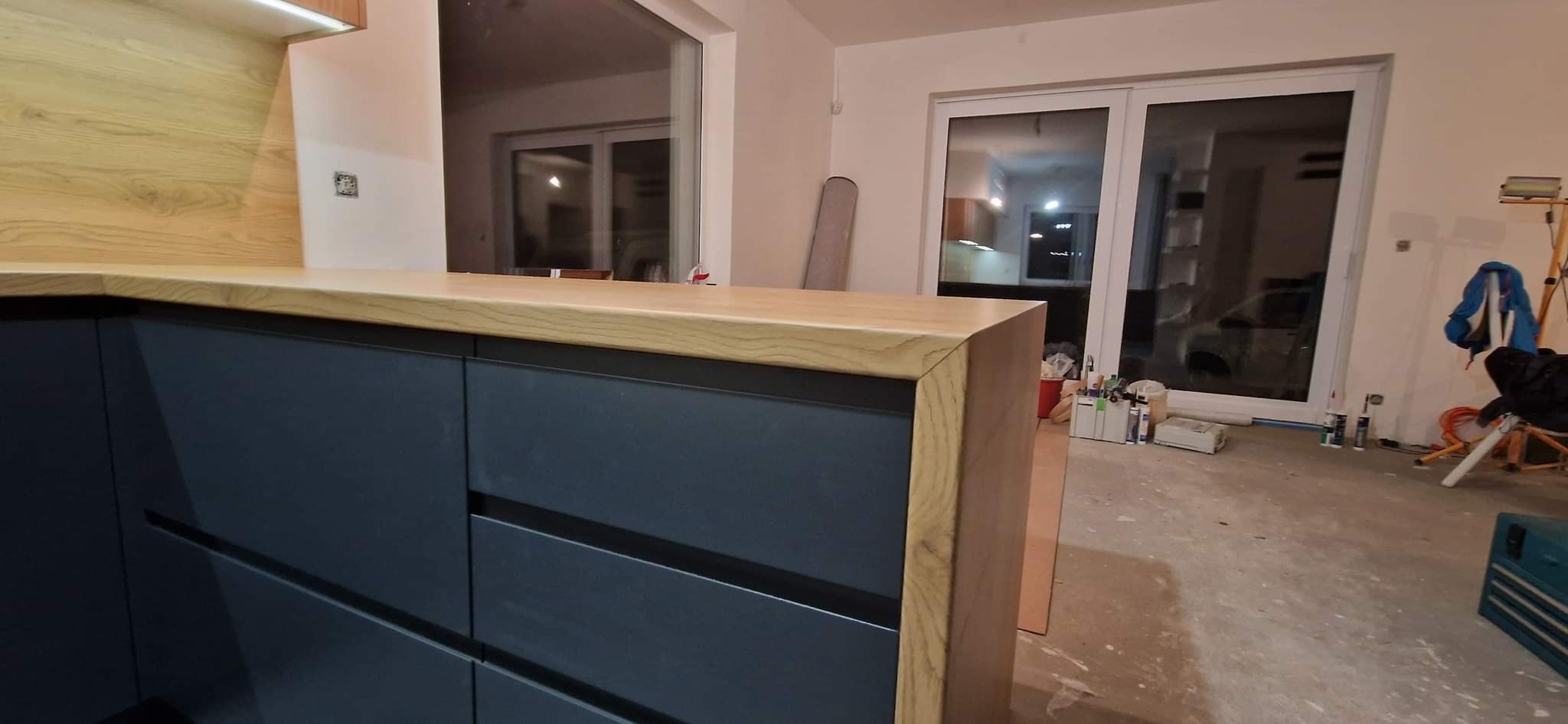 Ciemnoniebieskie szafki kuchenne z drewnianym blatem, widoczne materiały budowlane i narzędzia na podłodze w tle.