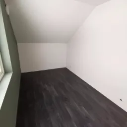 Poddasze po remoncie: świeżo pomalowane białe ściany, ciemna podłoga z paneli imitujących drewno, fragment ściany przy oknie pomalowany na oliwkowy kolor, widoczne punkty oświetleniowe w podłodze.