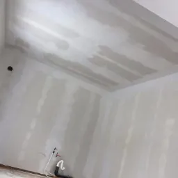 Pokój w trakcie prac przygotowawczych do malowania, ściany i sufit pokryte nierównomiernie nałożoną warstwą gładzi szpachlowej, widoczne rury przy podłodze, kubeł z farbą w rogu pomieszczenia.