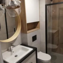 Nowoczesna łazienka z umywalką nablatową, wiszącą toaletą i kabiną prysznicową z czarnymi profilami, wykończona płytkami w odcieniach szarości i drewna, z okrągłym lustrem i lampą w folii ochronnej.