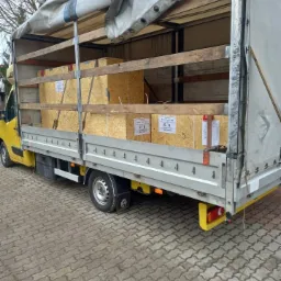 Żółty bus z plandeką załadowany drewnianymi skrzyniami z etykietami, widok z boku na brukowanym podłożu.