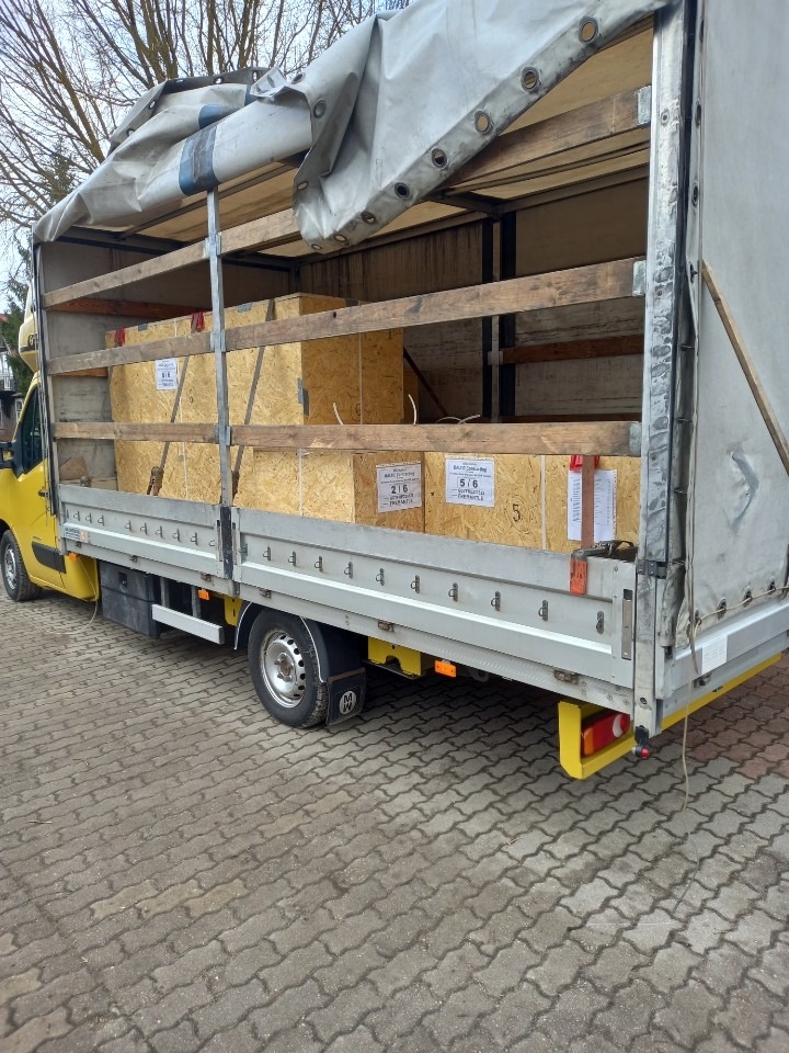 Żółty bus z plandeką załadowany drewnianymi skrzyniami z etykietami, widok z boku na brukowanym podłożu.