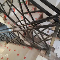 Metalowa balustrada schodowa o geometrycznym wzorze, czarna, montowana na schodach w trakcie wykańczania, widoczne zabezpieczenia na stopniach i narzędzia pracy.